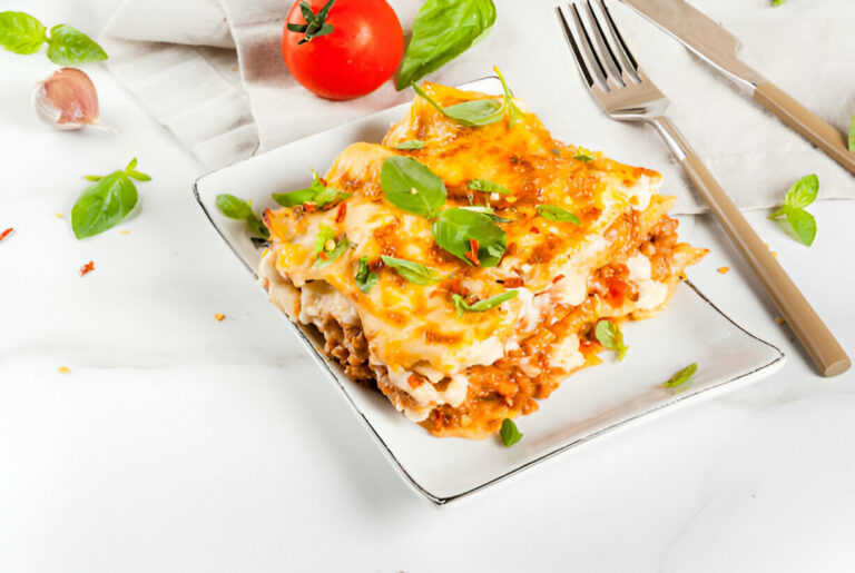 A delicious serving of lasagna topped with melted cheese and fresh basil, accompanied by a tomato and garlic on a marble surface.