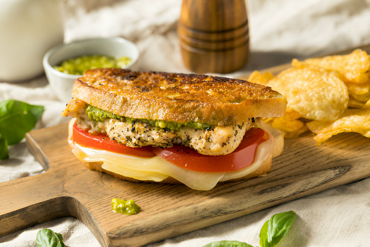 A grilled sandwich with chicken, tomato, mozzarella cheese, and pesto on a wooden cutting board, accompanied by potato chips and a small bowl of green sauce.