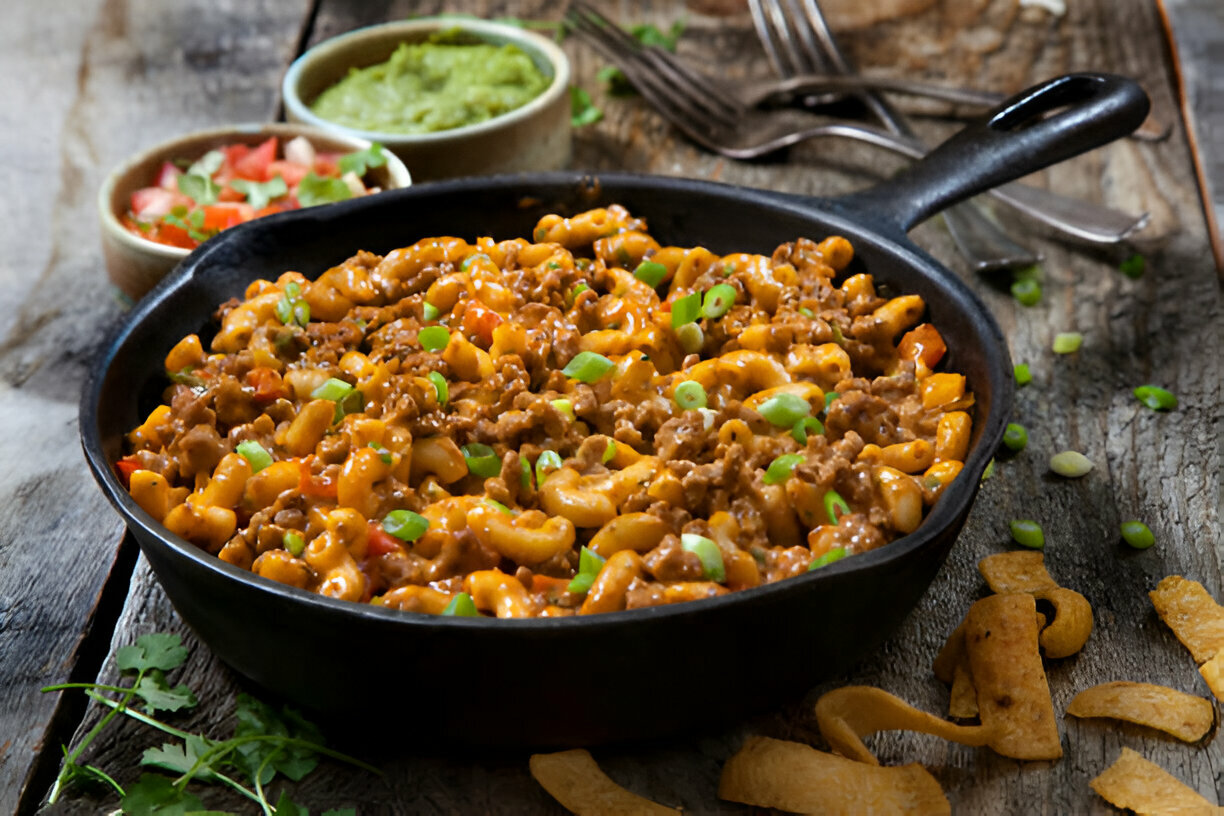 A cast iron skillet filled with macaroni, ground beef, and green onions, served with guacamole and salsa on the side.