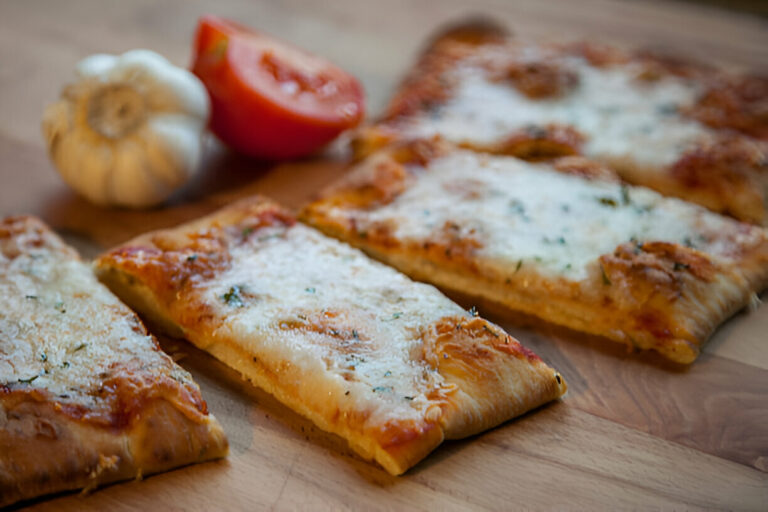 Four slices of cheesy pizza on a wooden surface alongside garlic and a tomato.