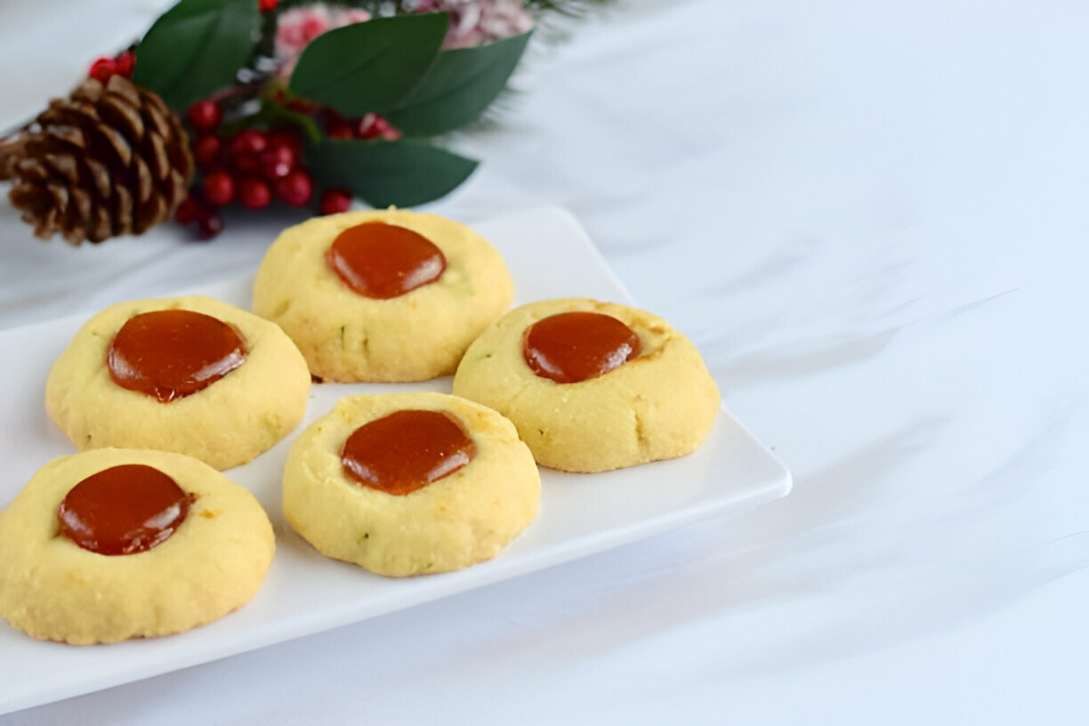 A plate of five soft cookies topped with a shiny jam-like glaze, set against a white background with decorative pine and berries.