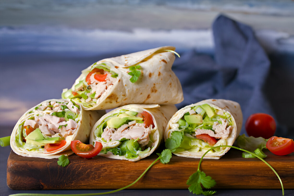 Four wraps filled with chicken, lettuce, cucumbers, and tomatoes placed on a wooden cutting board.