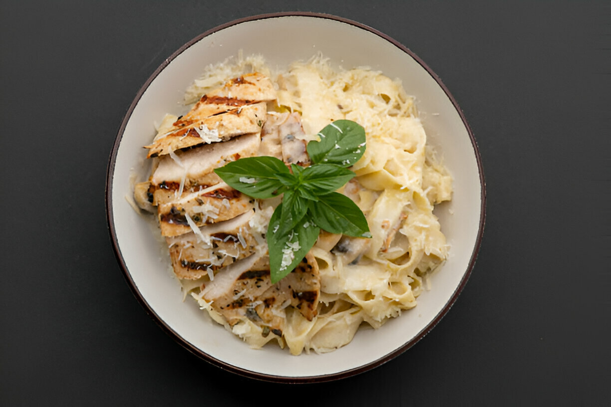 A bowl of fettuccine Alfredo topped with grilled chicken and fresh basil leaves