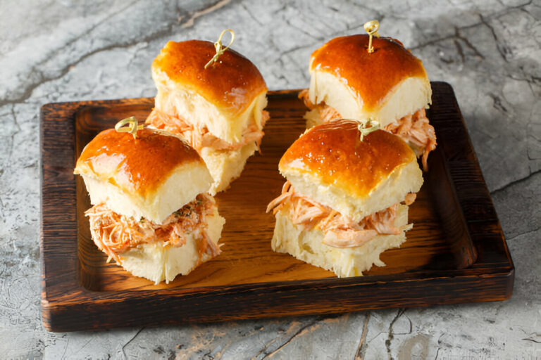 A wooden platter with four pulled chicken sliders, each topped with a shiny, golden-brown bun.