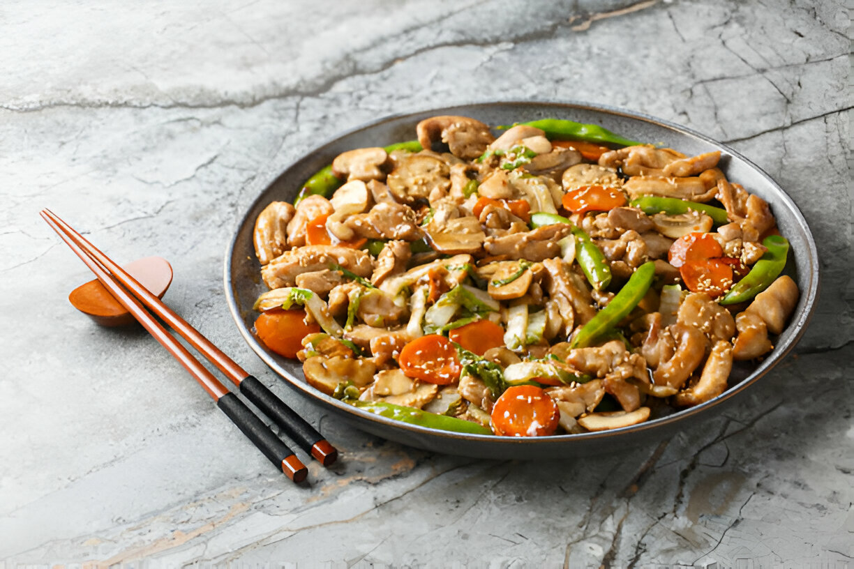 A bowl of stir-fried chicken with vegetables, including green bell peppers, carrots, and sesame seeds, served with chopsticks.