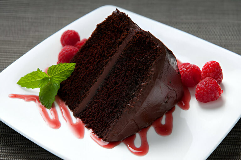 A slice of chocolate cake garnished with fresh raspberries and mint leaves on a white plate.