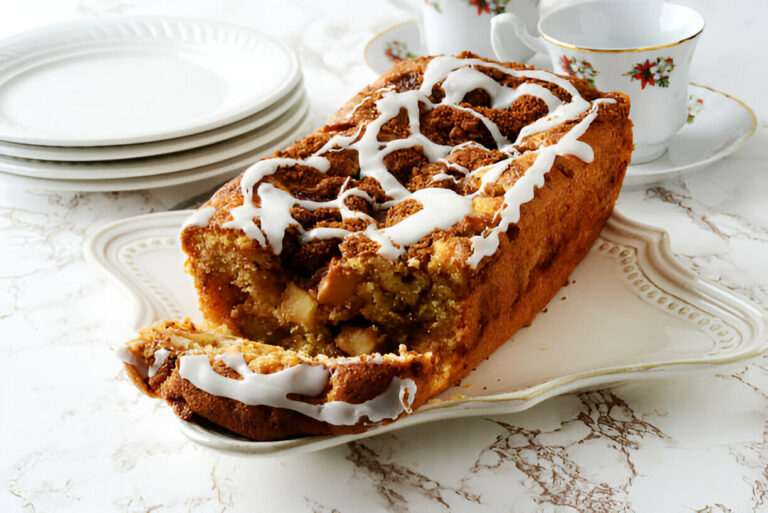 A sliced loaf cake with icing, revealing apple filling inside, served on a decorative plate with stacked white plates and teacups in the background.