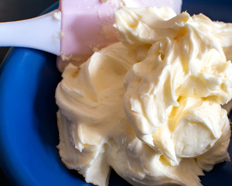 A close-up image of creamy butter or frosting in a blue bowl with a pink spatula partially visible.