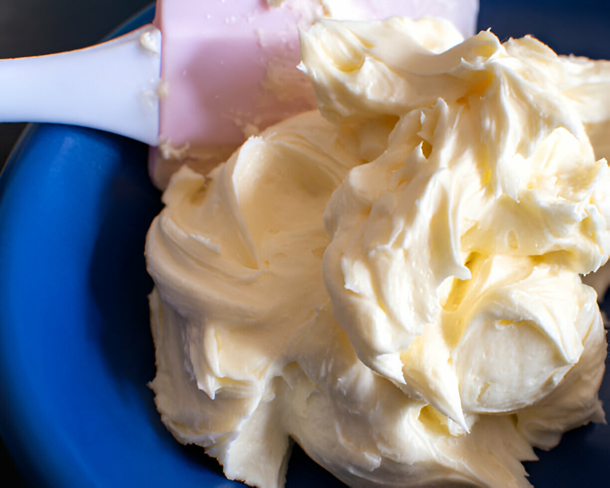 A close-up image of creamy butter or frosting in a blue bowl with a pink spatula partially visible.