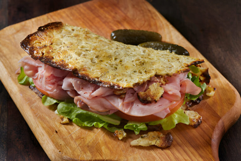 A delicious sandwich with layers of ham, lettuce, and tomato, topped with a golden, crispy crust, served on a wooden cutting board alongside pickles.