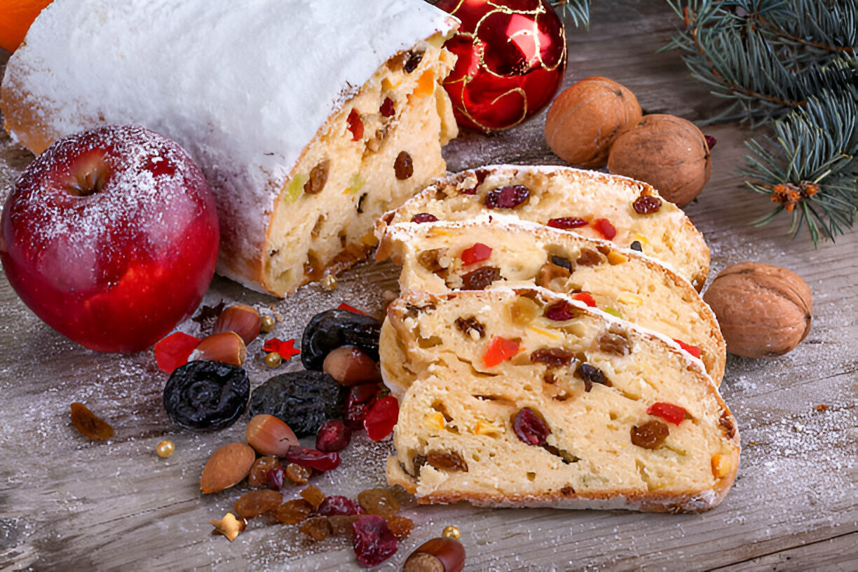 A festive spread featuring a sliced loaf of stollen bread dusted with powdered sugar, surrounded by an apple, dried fruits, nuts, and Christmas decorations.