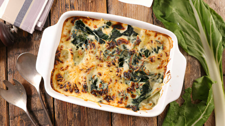 A white dish filled with baked greens and cheese resting on a wooden table, alongside some fresh leafy greens.