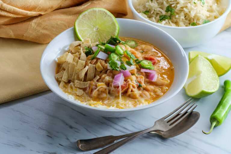 A bowl of comforting bean soup topped with green onions, pink onions, and tortilla strips, served with rice and lime wedges.