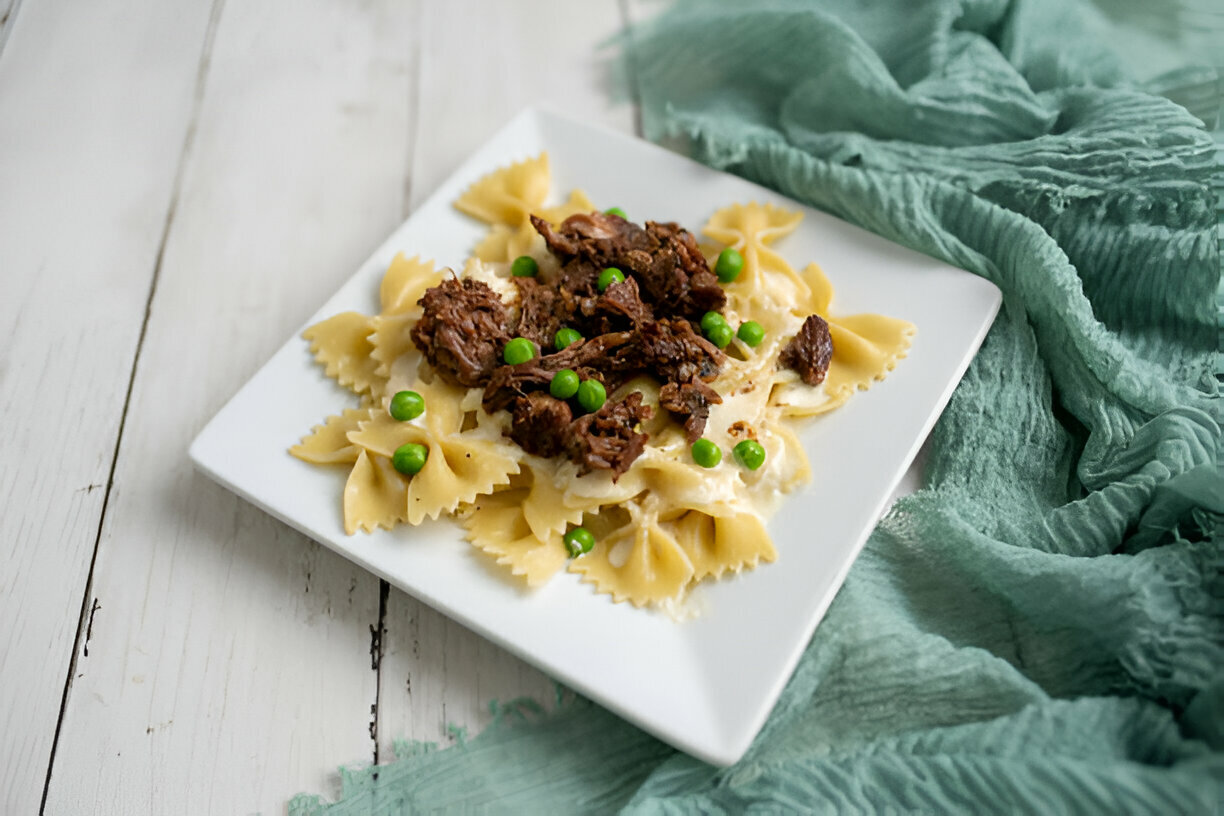 A white plate with farfalle pasta topped with shredded beef and green peas, set on a rustic wooden table.