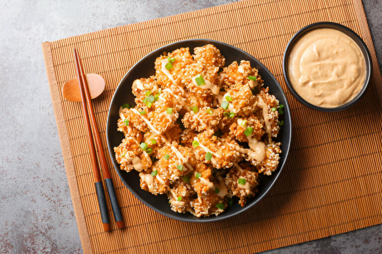 A black bowl filled with crispy popcorn chicken topped with green onions and drizzled with sauce, served on a bamboo mat with a small bowl of dipping sauce.