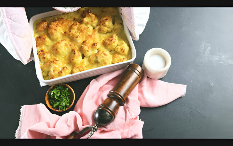 Baked cauliflower dish with a golden topping in a white baking dish, surrounded by fresh herbs, a pink cloth, and pepper grinder.