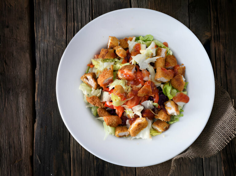 A fresh salad topped with crispy chicken pieces, drizzled with dressing, served in a white bowl on a rustic wooden table.