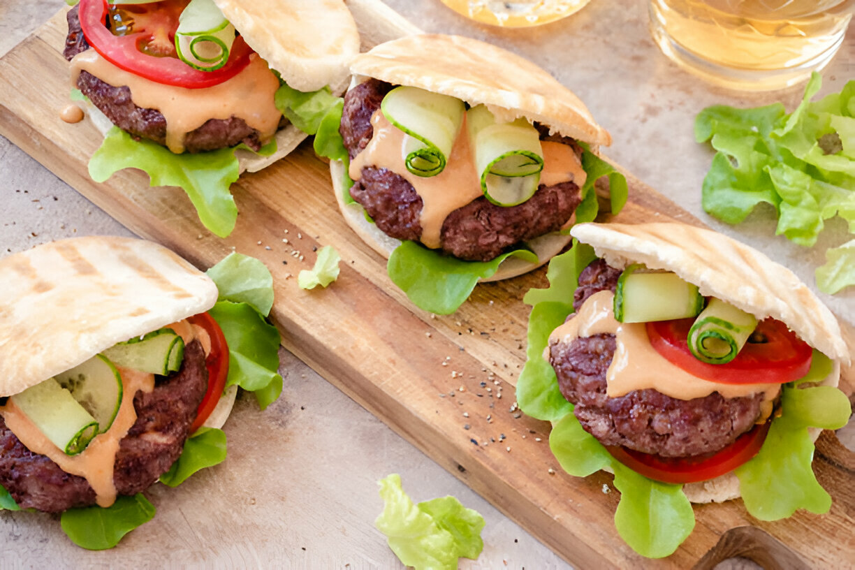 Three mini burgers with lettuce, tomatoes, cucumbers, and sauce on a wooden board