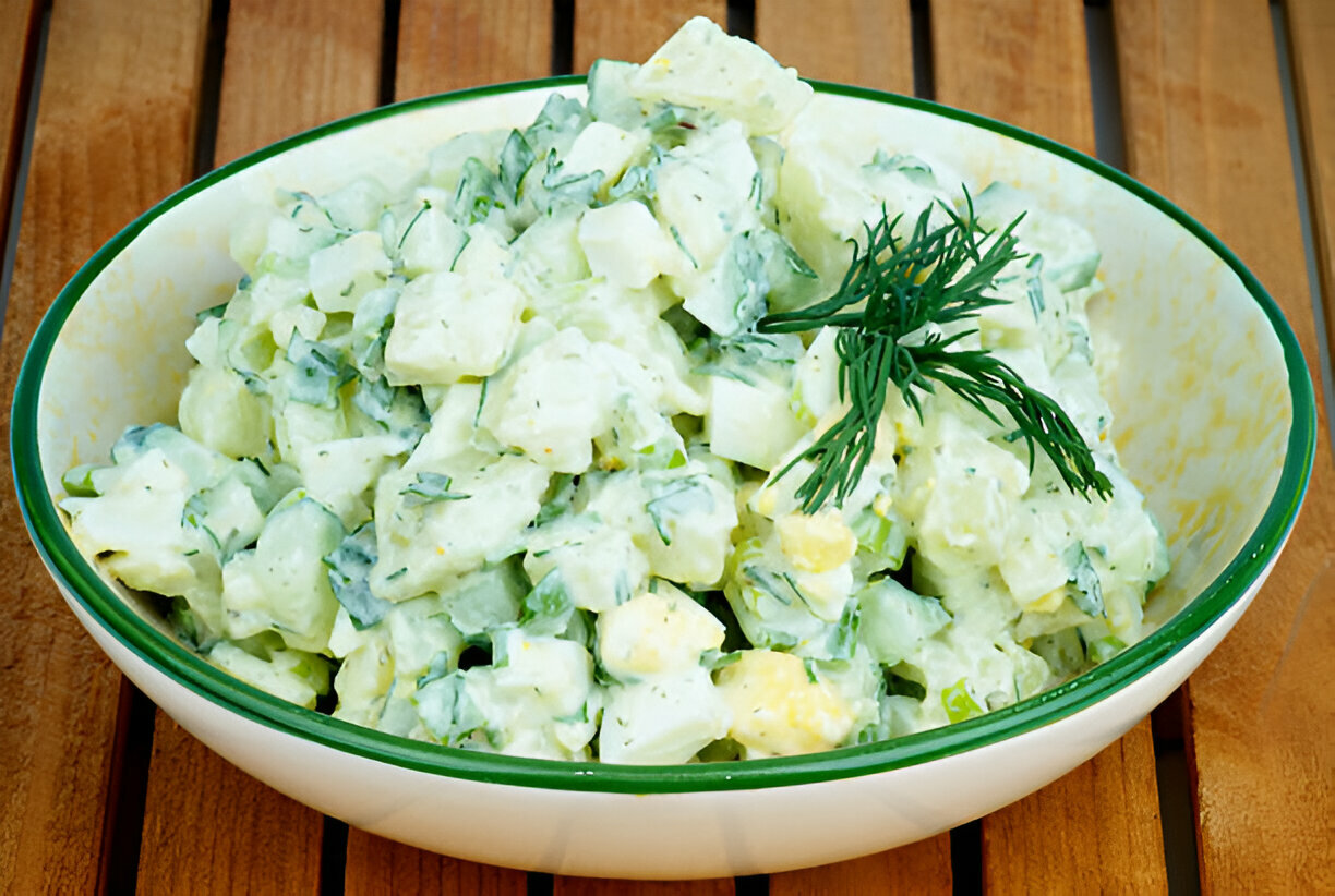 A bowl of potato salad with diced cucumbers and fresh dill on a wooden table.