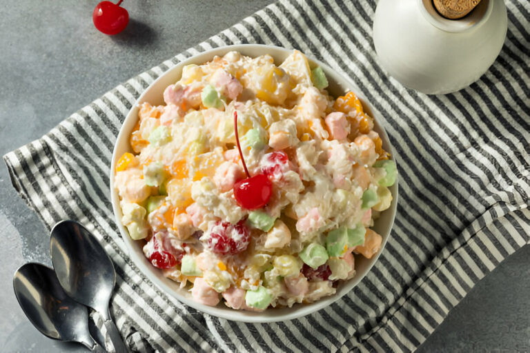 A colorful fruit salad in a white bowl topped with a cherry, sitting on a striped cloth.