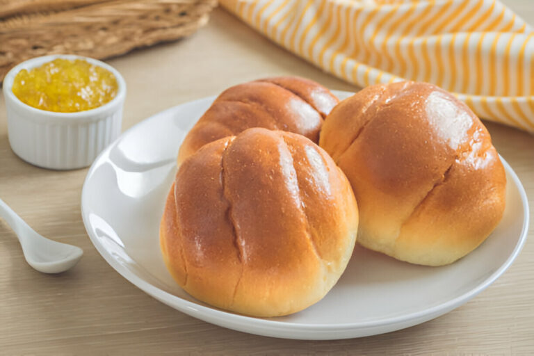 Three freshly baked golden brown bread rolls on a white plate next to a small dish of yellow jelly.