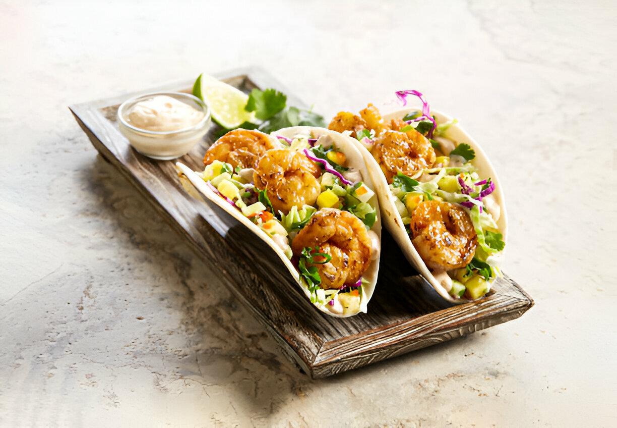 Three shrimp tacos with cabbage and mango salsa on a wooden serving board, accompanied by a lime wedge and dipping sauce.