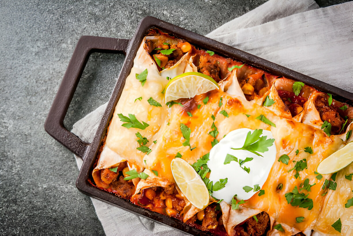 Baked enchiladas topped with cheese, sour cream, cilantro, and lime slices in a baking dish.