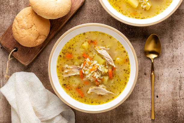 A bowl of chicken soup with vegetables and rice, accompanied by two rolls on a wooden board.