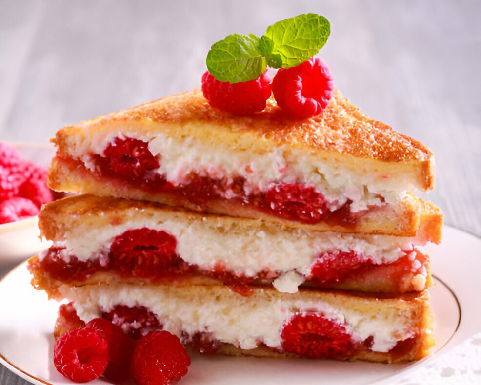 A stack of golden-brown grilled cheese sandwiches filled with raspberries and cream cheese, garnished with fresh mint.