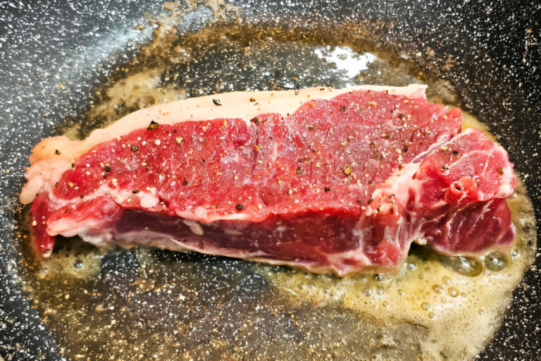 A thick cut of steak being cooked in a frying pan with sizzling butter and spices.