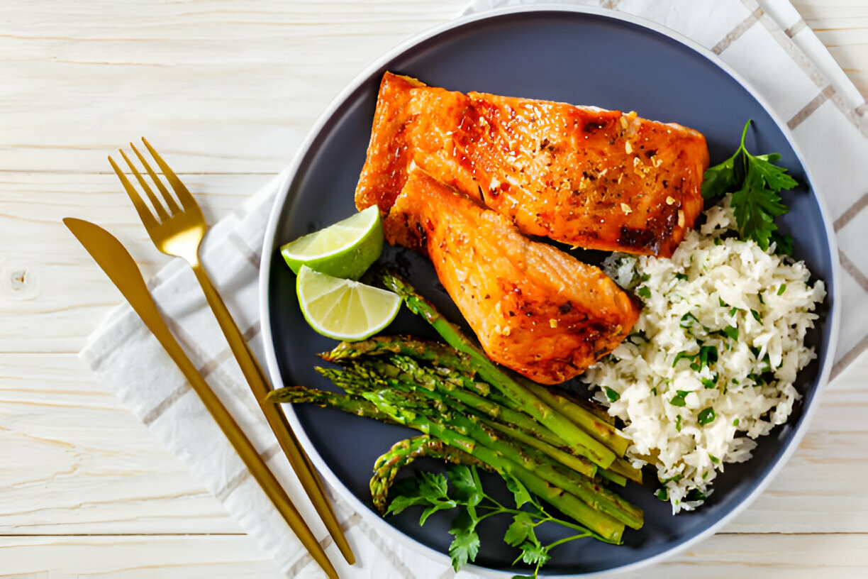 A plated meal featuring grilled salmon, asparagus, rice, and lime wedges on a dark plate