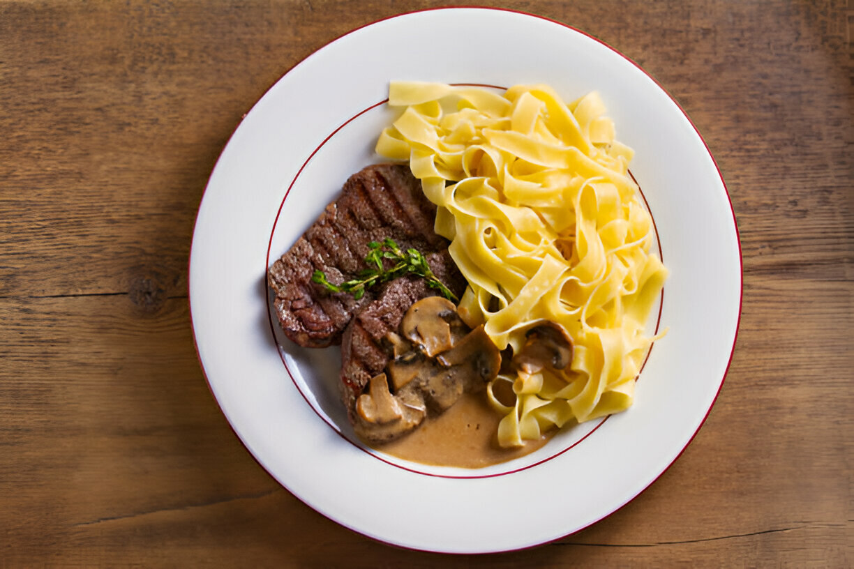 A plate of cooked steak with mushrooms in sauce alongside fettuccine pasta arranged neatly on a wooden table.