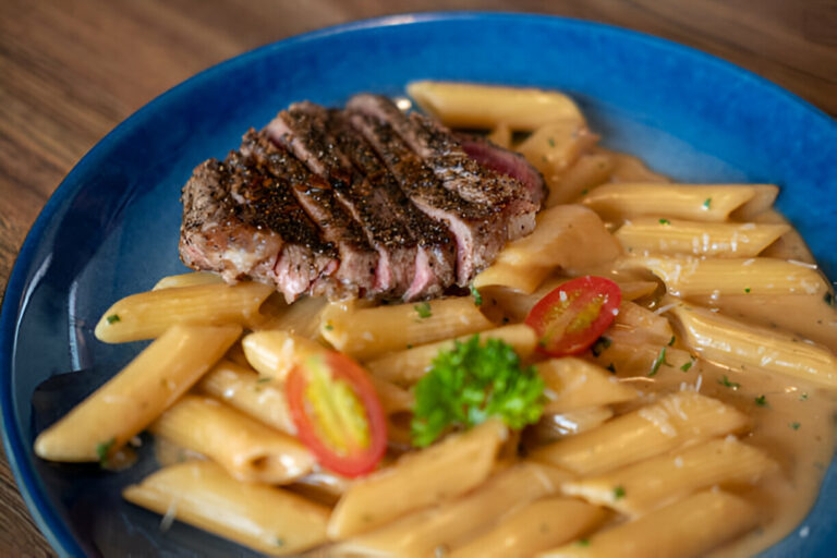 A plate of creamy penne pasta topped with a grilled steak and cherry tomatoes.