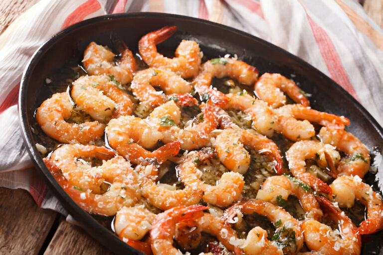 A close-up view of baked shrimp with a golden crust, garnished with herbs, served in a black dish on a rustic wooden table.