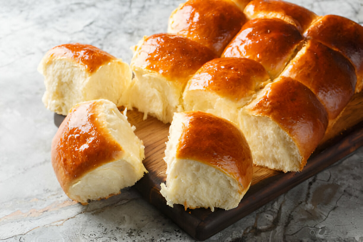 Freshly baked golden brown rolls arranged on a wooden board, with a few pieces cut and displayed beside them.