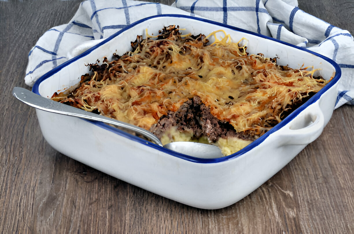 A baked casserole dish with a golden brown cheesy topping and a portion removed, revealing layers of ground meat and potatoes.