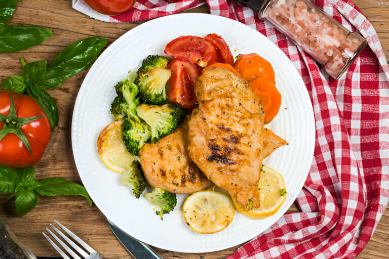 Grilled chicken breast served with broccoli, tomatoes, and lemon slices on a white plate with a checkered cloth in the background.