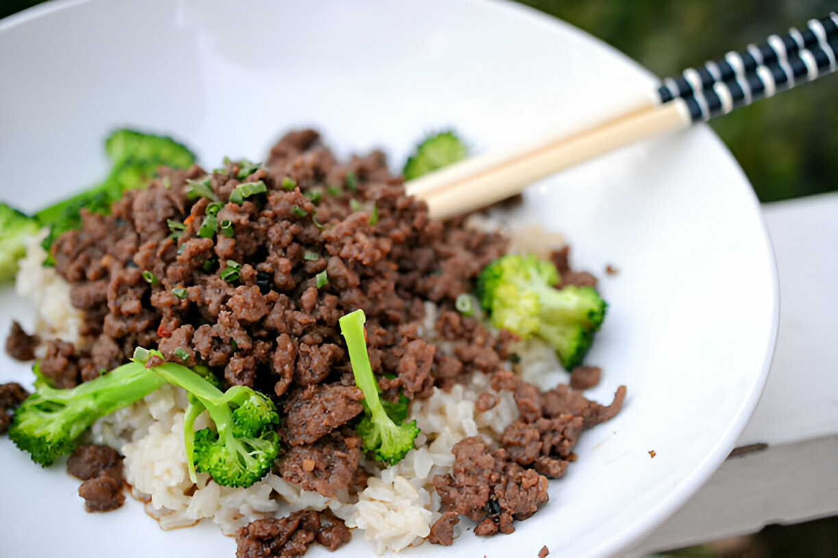 A bowl of cooked rice topped with finely minced beef and fresh broccoli, with chopsticks resting on the side.