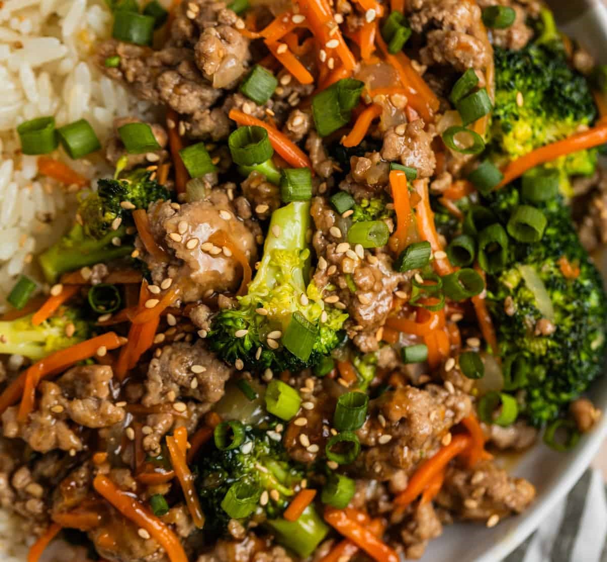 A vibrant bowl of stir-fried beef with broccoli, carrots, and green onions served alongside white rice, topped with sesame seeds.