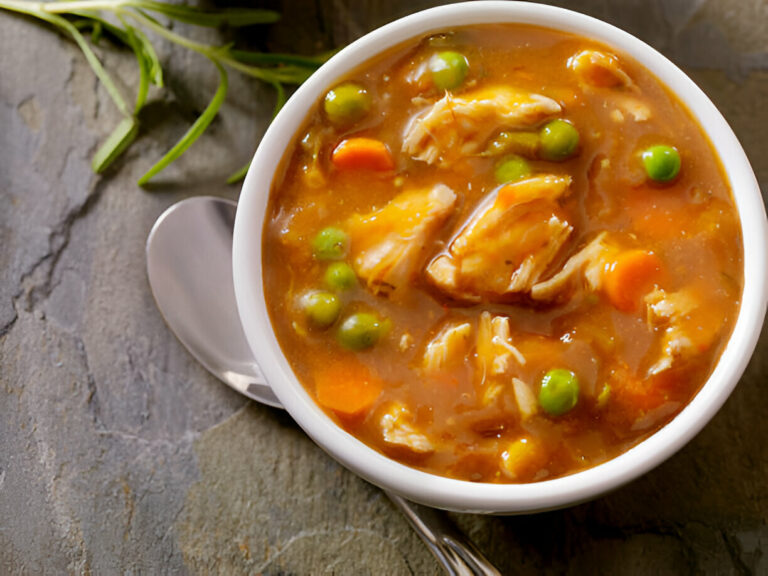 A bowl of chicken and vegetable soup with carrots and peas on a textured surface.