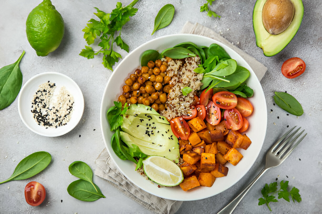 A colorful vegan bowl filled with quinoa, chickpeas, fresh greens, cherry tomatoes, sweet potatoes, and garnished with lime and sesame seeds.