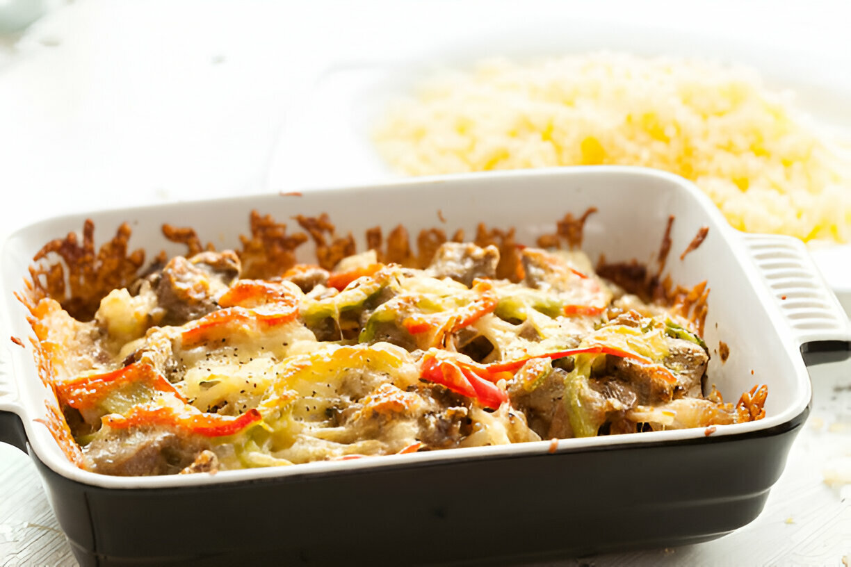 A delicious baked dish with meat and colorful vegetables topped with melted cheese in a black and white baking dish, accompanied by a serving of rice in the background.