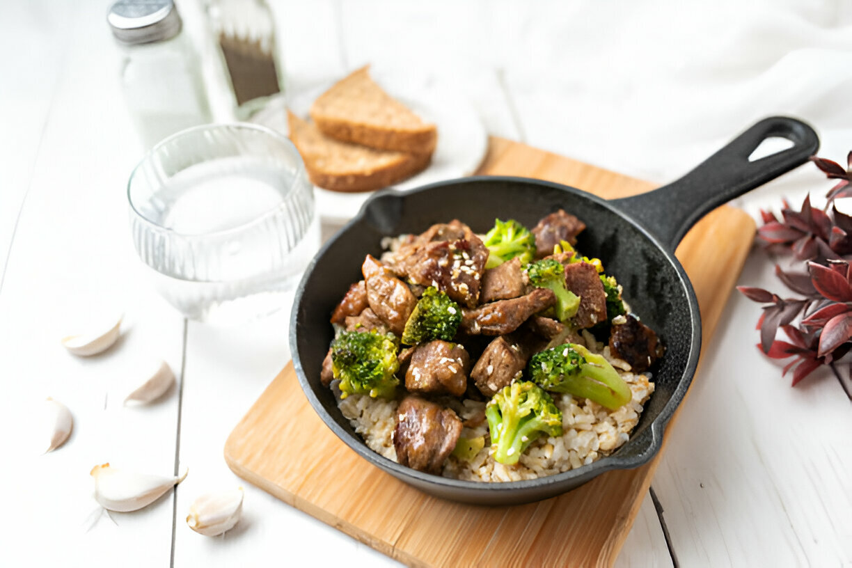 A skillet of stir-fried beef and broccoli served over rice, accompanied by a glass of water and slices of brown bread in the background.