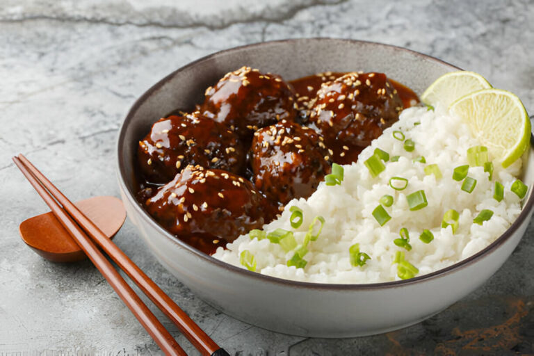 A bowl of meatballs in a savory sauce served with fluffy white rice and garnished with green onions and lime slices.