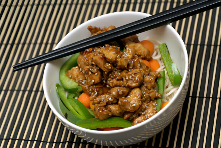 A bowl of chicken stir-fry with vegetables served over noodles, topped with sesame seeds and chopsticks resting on the bowl.