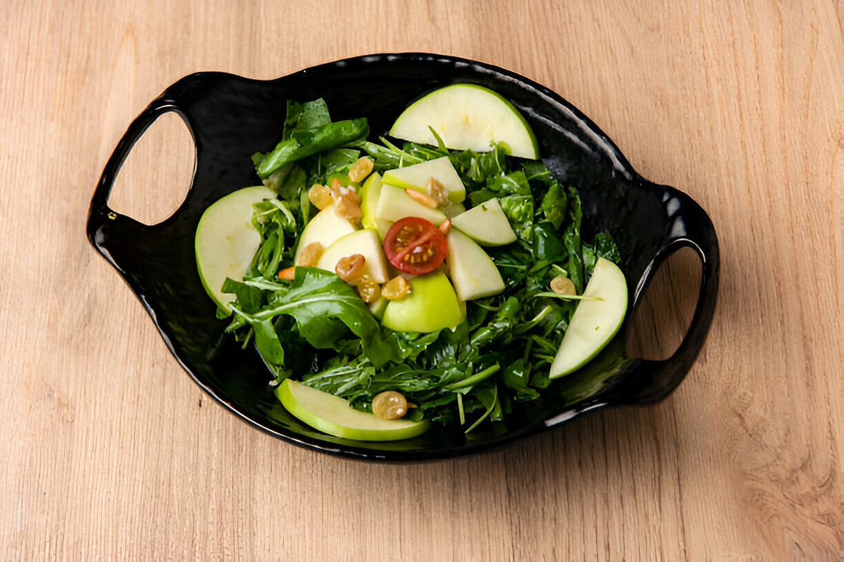 A fresh green salad with apple slices, cherry tomatoes, and nuts served in a black dish on a wooden surface.