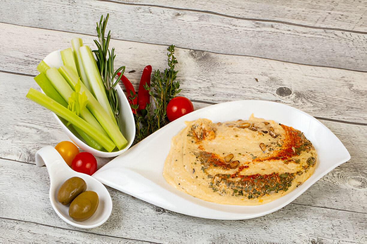 A white plate with hummus sprinkled with herbs, surrounded by fresh vegetables including celery, tomatoes, and olives.