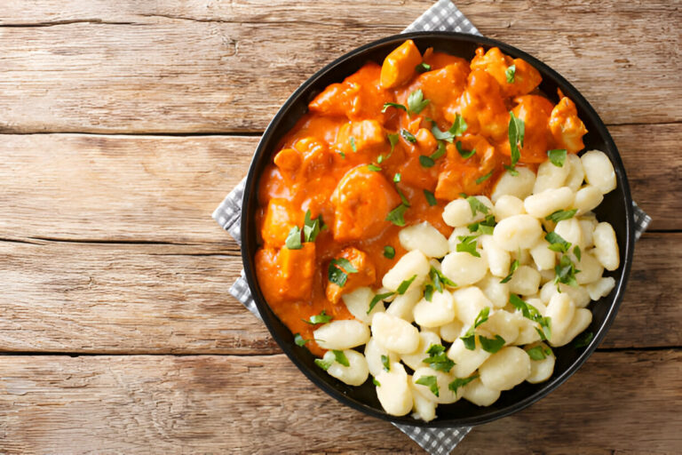 A black plate filled with chicken in a creamy sauce and gnocchi topped with fresh herbs, resting on a wooden surface.