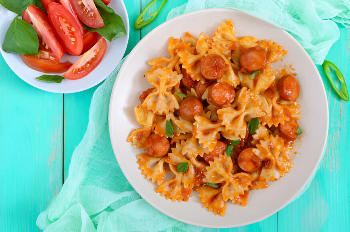 A bowl of farfalle pasta with sausage in tomato sauce, garnished with fresh basil leaves, served on a light green table.