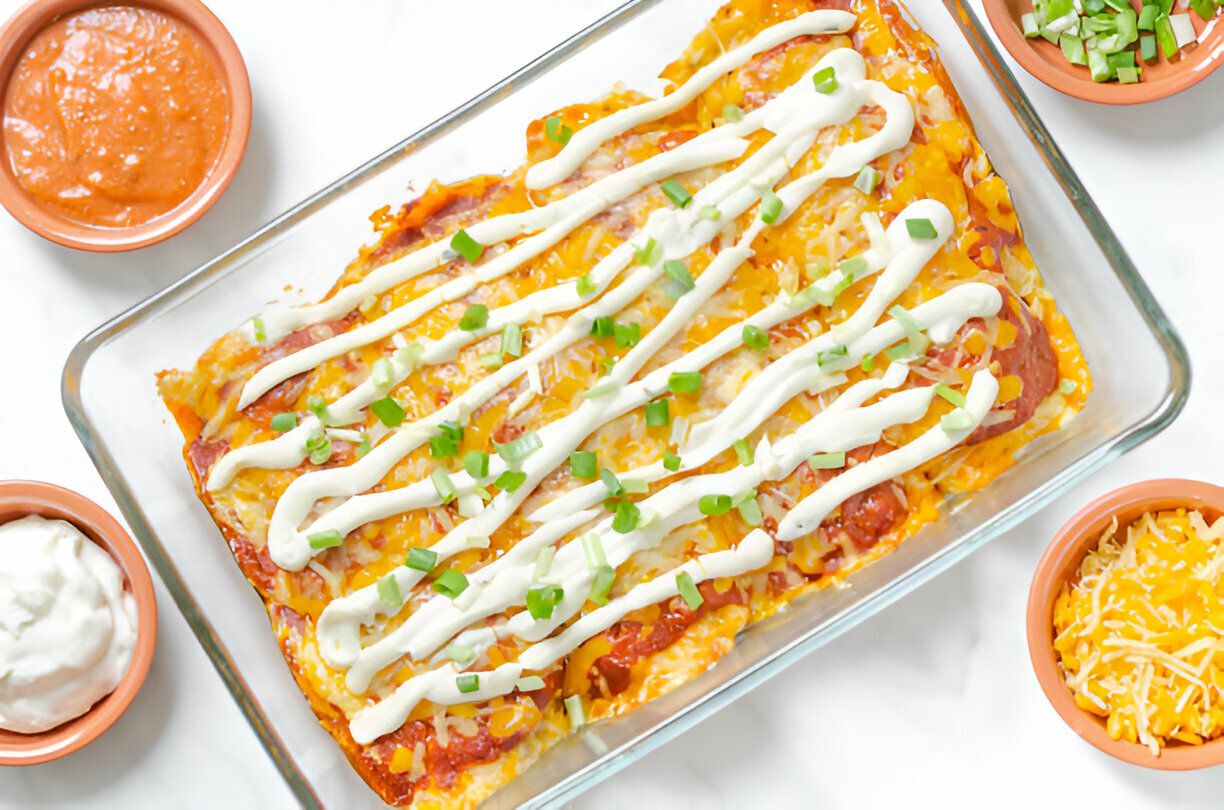 A delicious casserole dish of enchiladas topped with cheese and green onions, surrounded by small bowls of sauce and shredded cheese.